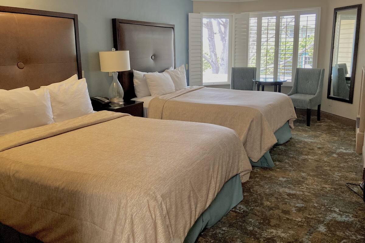 A hotel room with two beds, a nightstand, a lamp, a chair, and a large window with blinds.