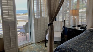 A hotel room with a bed, a desk with a lamp, a chair, and a view of the beach through open shutters. - Blue Dolphin Inn (Cambria)