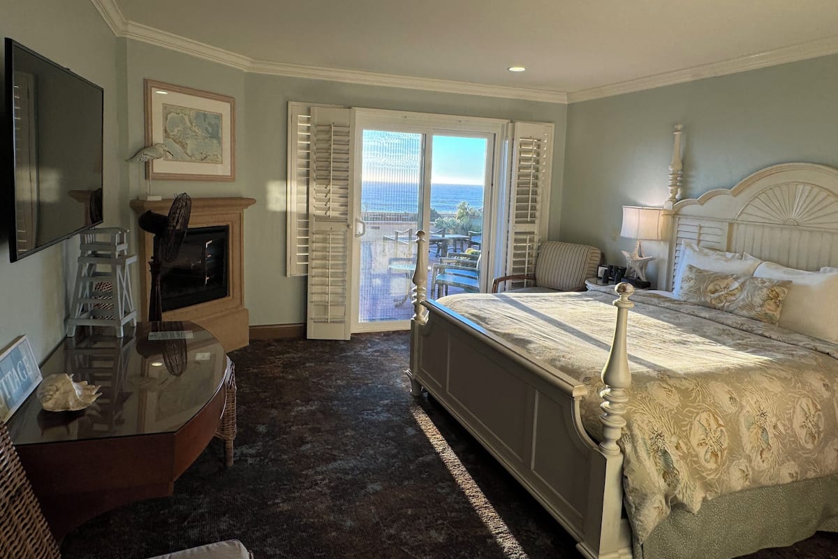 A bedroom with a large bed, a television, a fireplace, and a view of the ocean through open shutters.