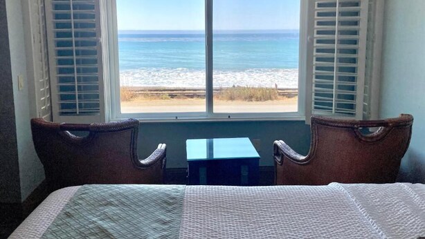A room with a bed, a small table, and a view of the beach through a window with shutters. - Blue Dolphin Inn (Cambria)