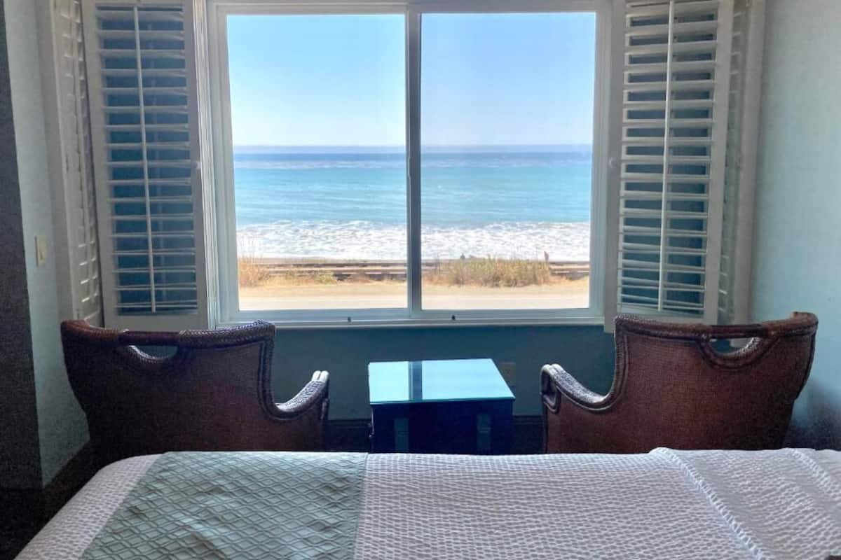 A room with a bed, a small table, and a view of the beach through a window with shutters.