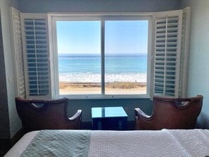 Una habitación con cama, una mesita y vista a la playa a través de una ventana con persianas.