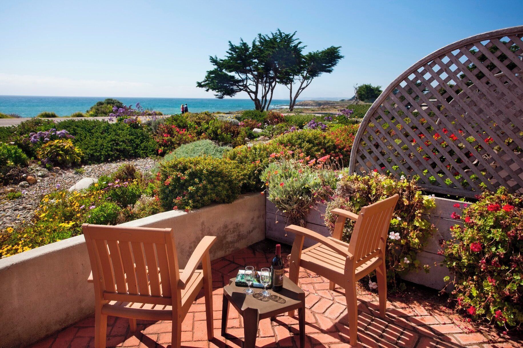 An outdoor seating area with two wooden chairs and a table, surrounded by a garden with various plants and flowers, overlooking a body of water.