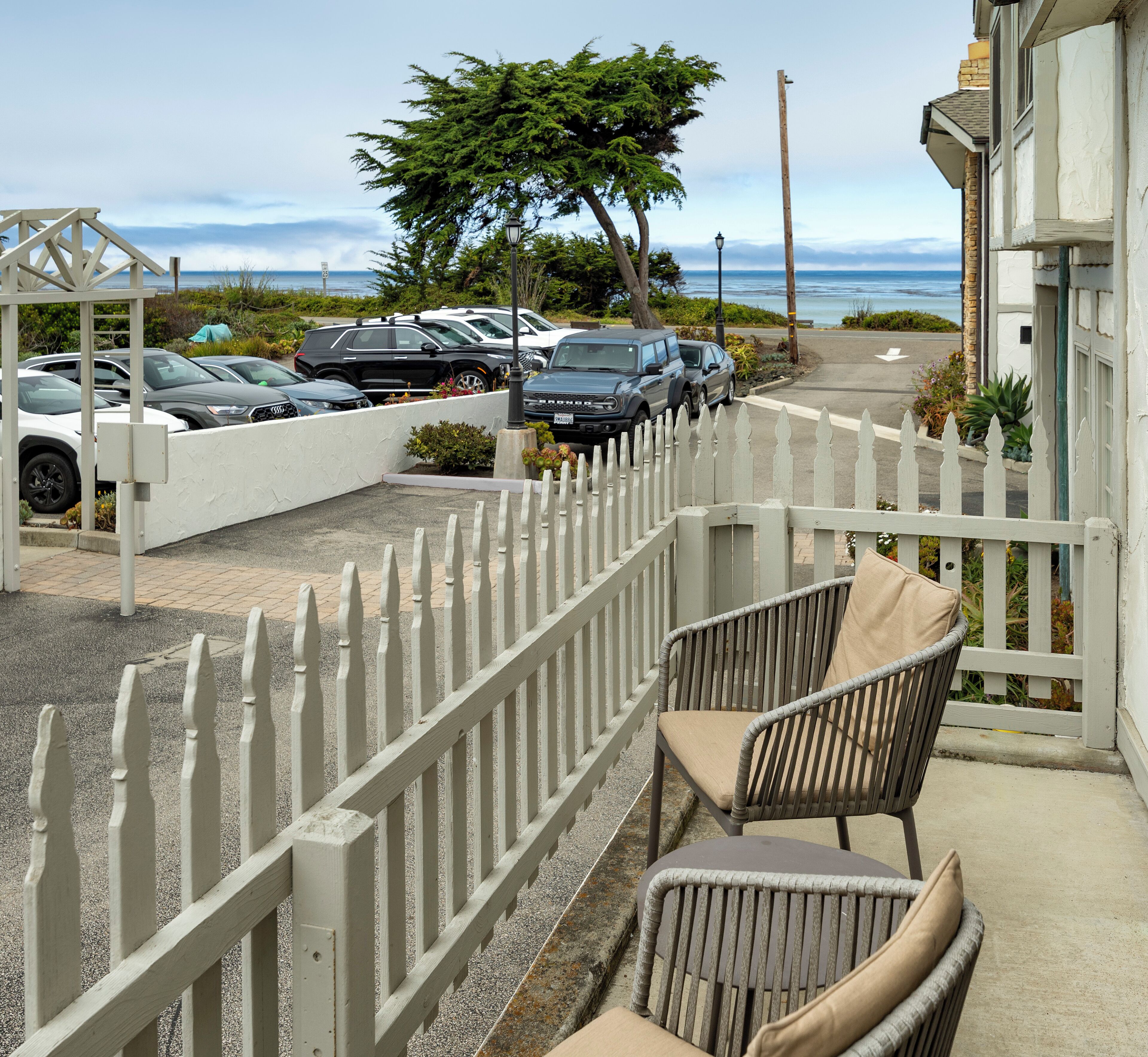 A balcony with a white picket fence, a wicker chair, and a table. In the background, there is a parking area with several cars and a view of the ocean.