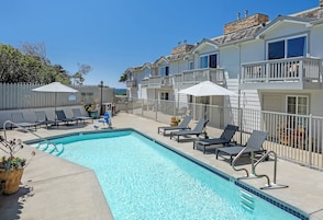 A swimming pool with lounge chairs and umbrellas, surrounded by a fence and a row of houses.