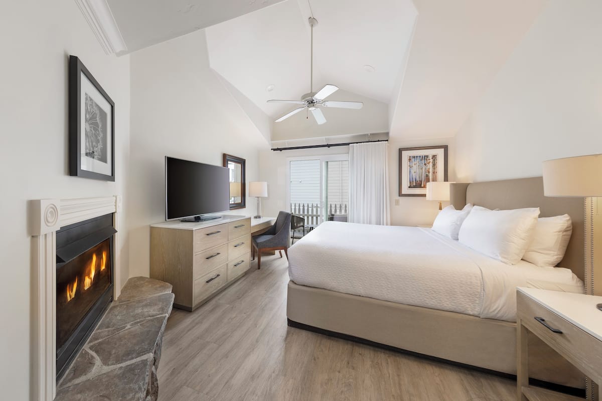 A hotel room with a fireplace, a bed, a desk with a television, and a balcony with curtains.