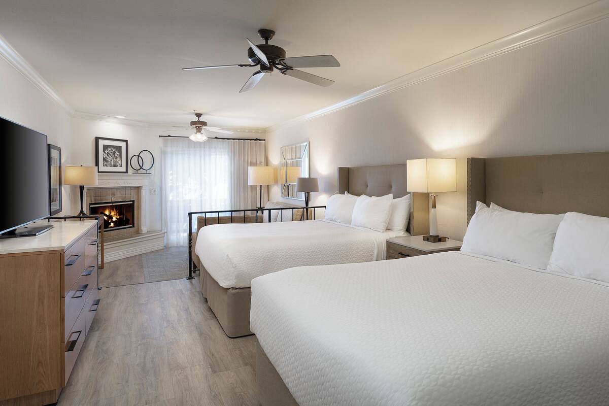 A hotel room with two beds, a fireplace, a ceiling fan, and a TV on a wooden dresser.