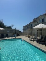 A clear swimming pool with lounge chairs and umbrellas, surrounded by a fenced area with a multi-story building.