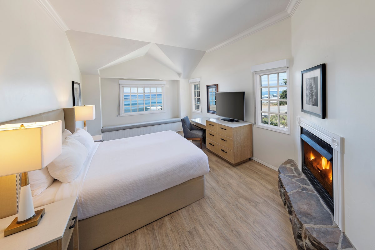 A bedroom with a bed, bedside table, lamp, TV, fireplace, and a view of the sea through the windows.