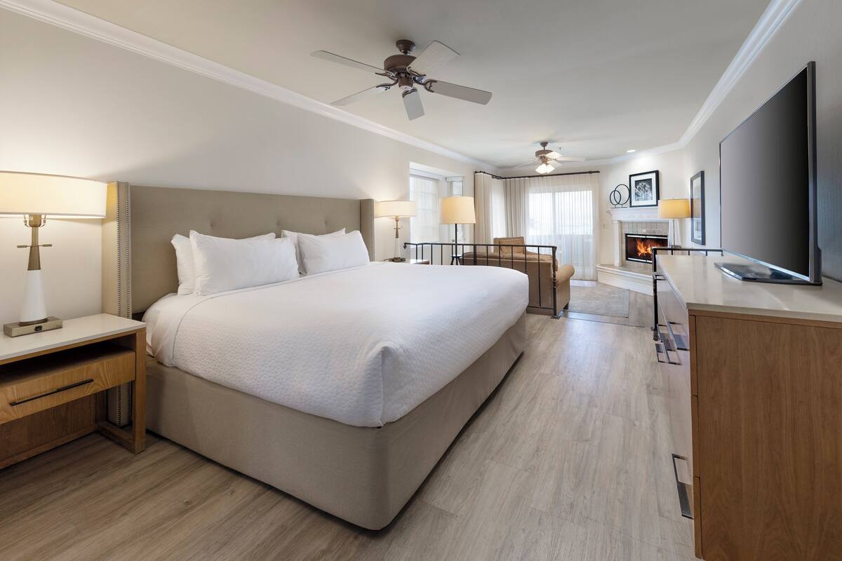 A hotel room with a large bed, a desk, a chair, a television, and a fireplace.