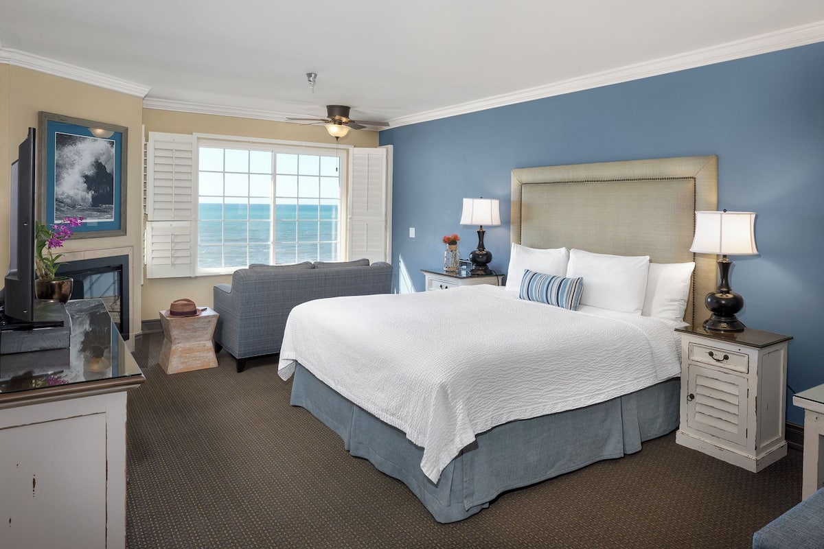 A hotel room with a large bed, a desk, a chair, a window with a view of the ocean, and a ceiling fan.