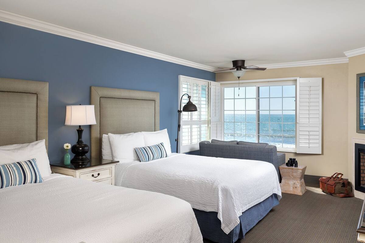 A hotel room with two beds, a nightstand with a lamp, a window with shutters, a ceiling fan, and a fireplace.