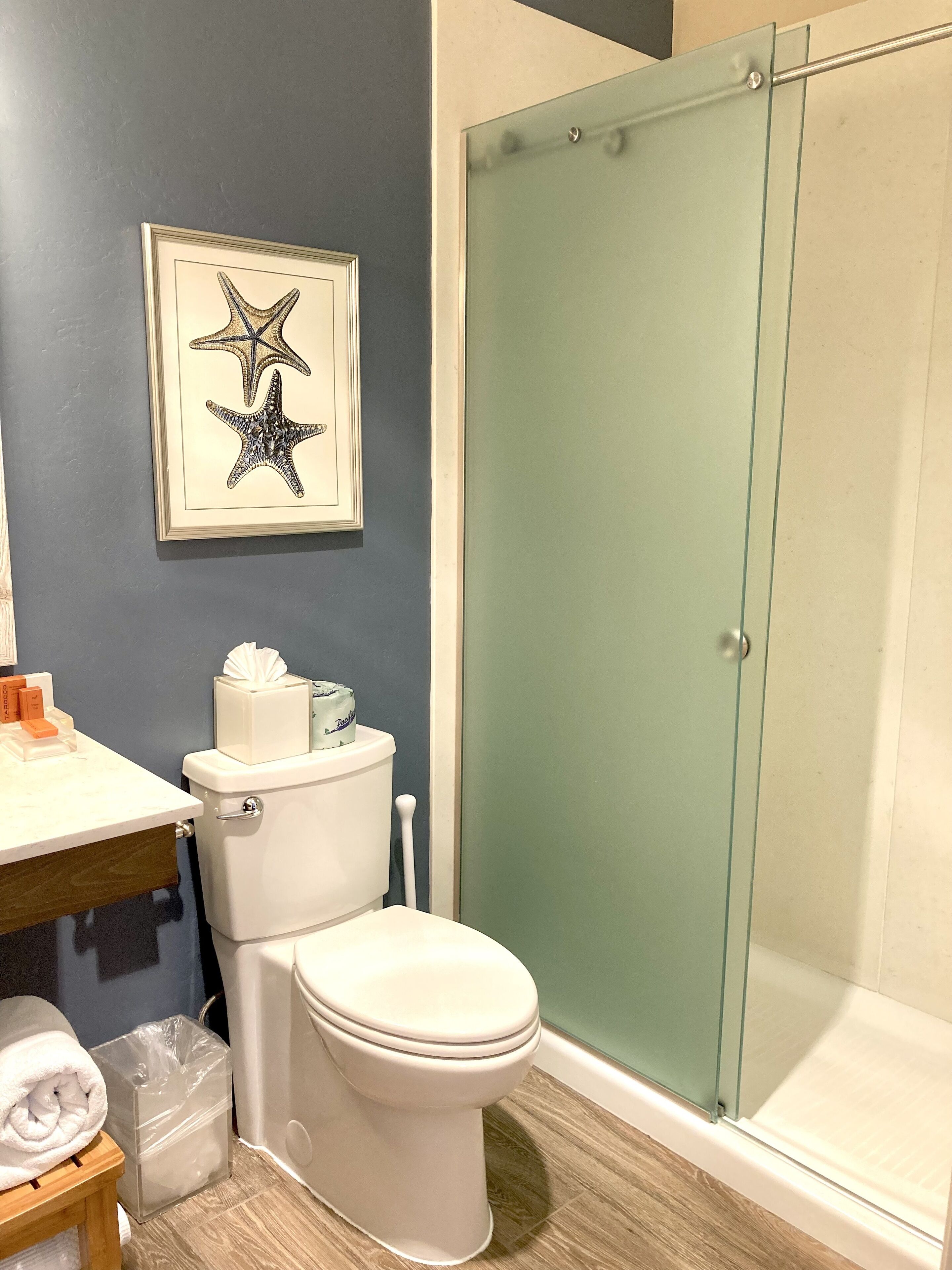 A bathroom with a toilet, a framed starfish artwork, a glass shower enclosure, and a wooden floor.