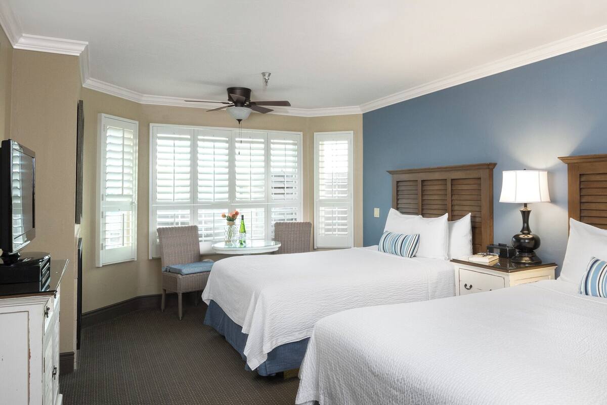 A hotel room with two beds, a ceiling fan, a TV, a small table with a vase, and a window with blinds.
