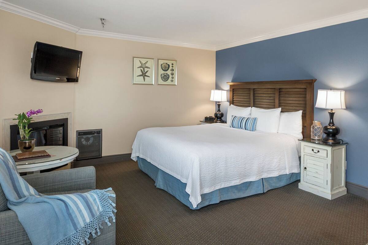 A hotel room with a large bed, two bedside tables with lamps, a TV mounted on the wall, a small round table with a vase of flowers, and a chair with a blue blanket.