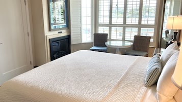A hotel room with a large bed, a small round table, two chairs, a fireplace, and a large window with blinds.