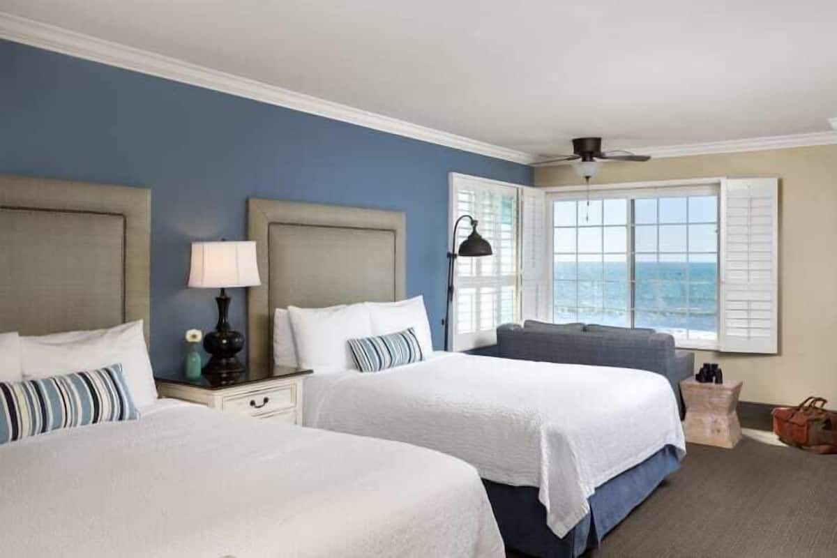 A hotel room with two beds, a large window with a view, a ceiling fan, and a nightstand with a lamp.