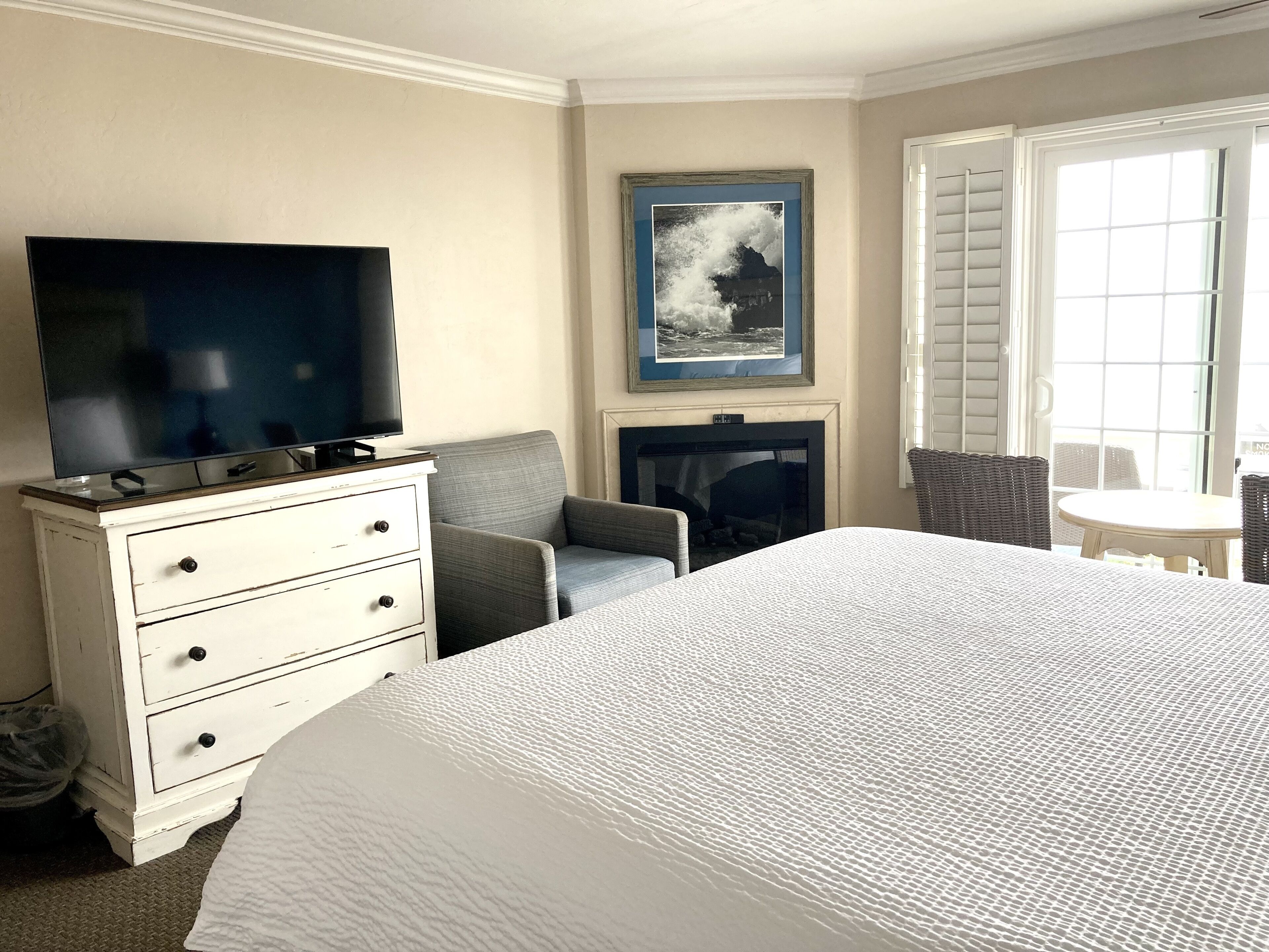 A hotel room with a large bed, a television on a dresser, a fireplace, a chair, and a window with blinds.