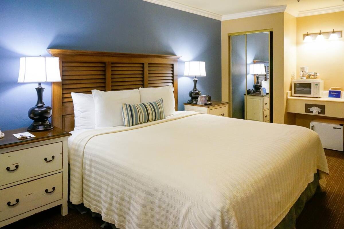 A neatly made bed with white linens and striped pillows, a wooden headboard, bedside tables with lamps, a small dresser, and a microwave in the corner.