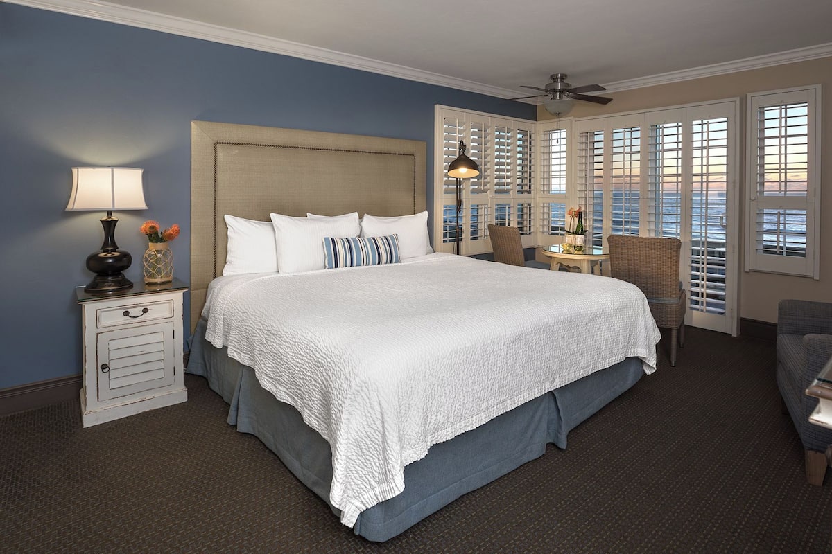 A large bed with a white quilt, a nightstand with a lamp, and a view of the ocean through a window with shutters.