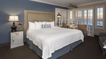A large bed with a white quilt, a nightstand with a lamp, and a view of the ocean through a window with shutters.