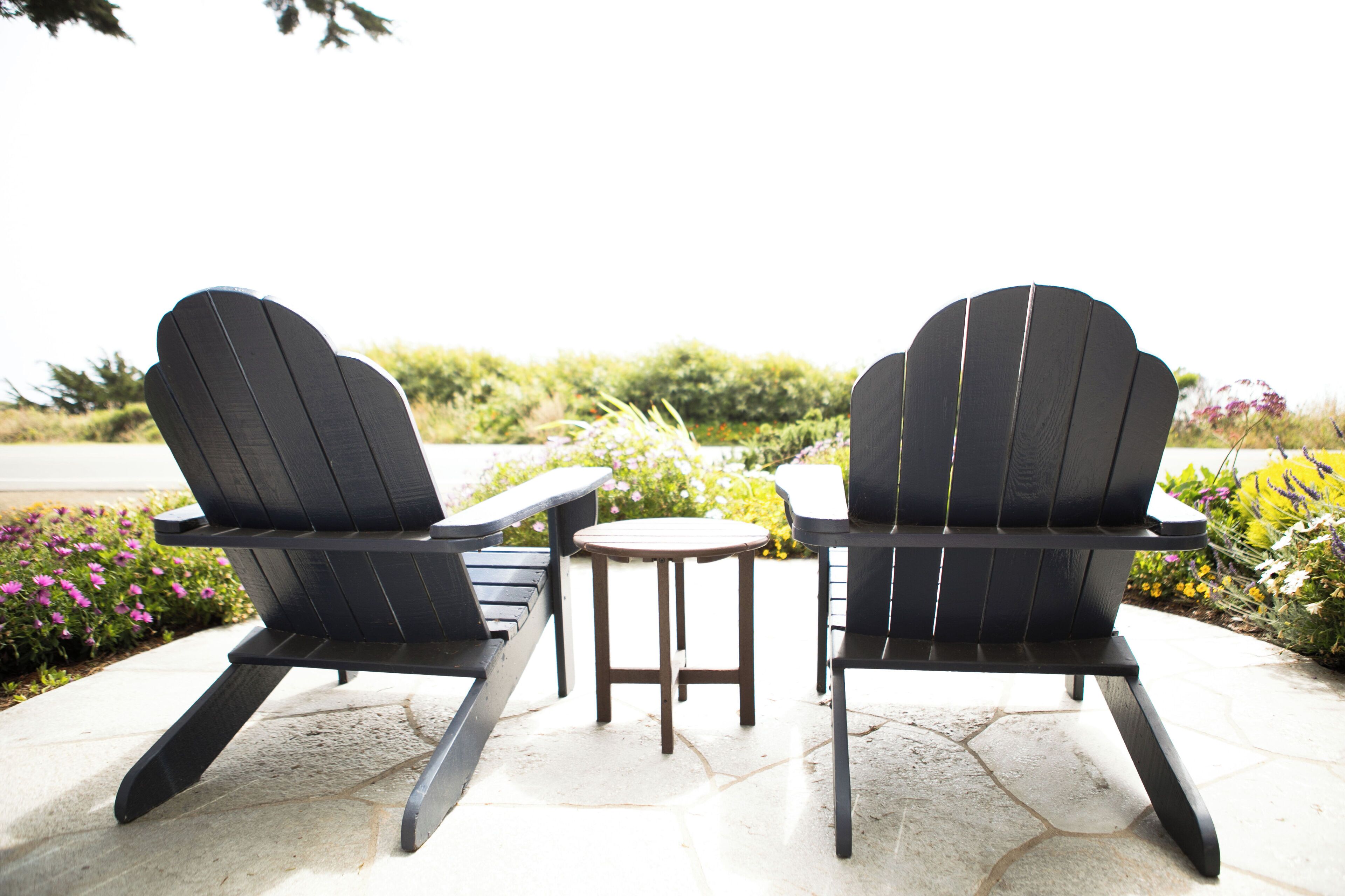 Two Adirondack chairs with a small round table between them, set on a stone patio with a garden in the background.