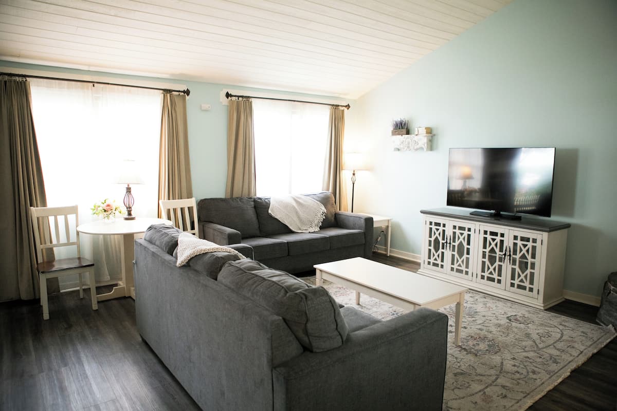 Ein Wohnzimmer mit grauem Sofa, weißem Couchtisch, Fernseher auf einem weißen Schrank und einem gemusterten Teppich.