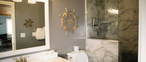 A bathroom with a large mirror, a ship's wheel decoration, a toilet, and a shower area with marble walls.