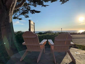 Two Adirondack chairs facing a scenic view of the ocean at sunset, with a wooden sign in the background. - Sea Otter Inn (Cambria)