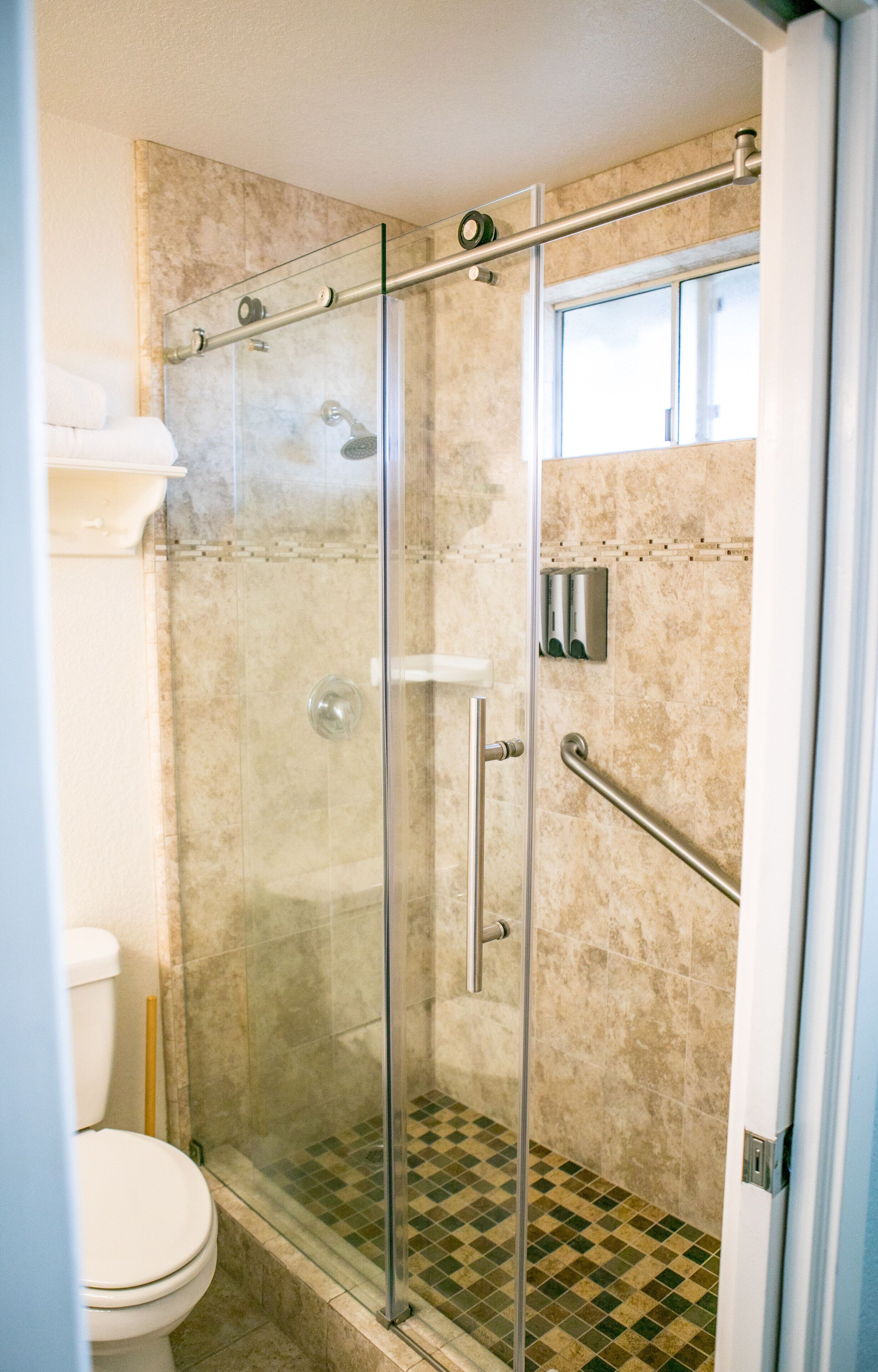 A glass-enclosed shower stall with a handrail, a toilet, and a checkered floor pattern.