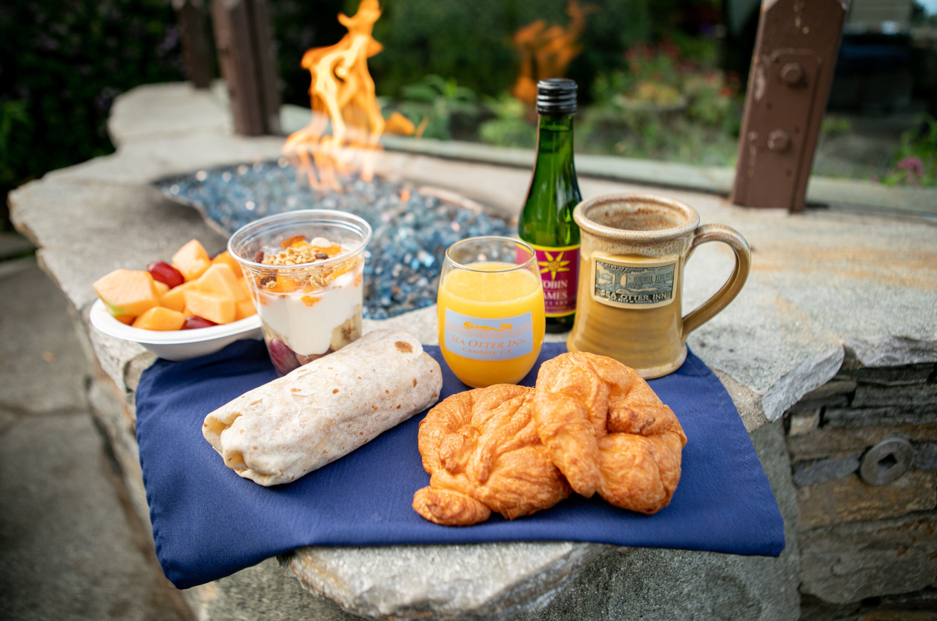 A breakfast setup with a stone table, a fire pit, a bottle of orange juice, a cup of coffee, a croissant, a wrap, and a bowl of fruit.