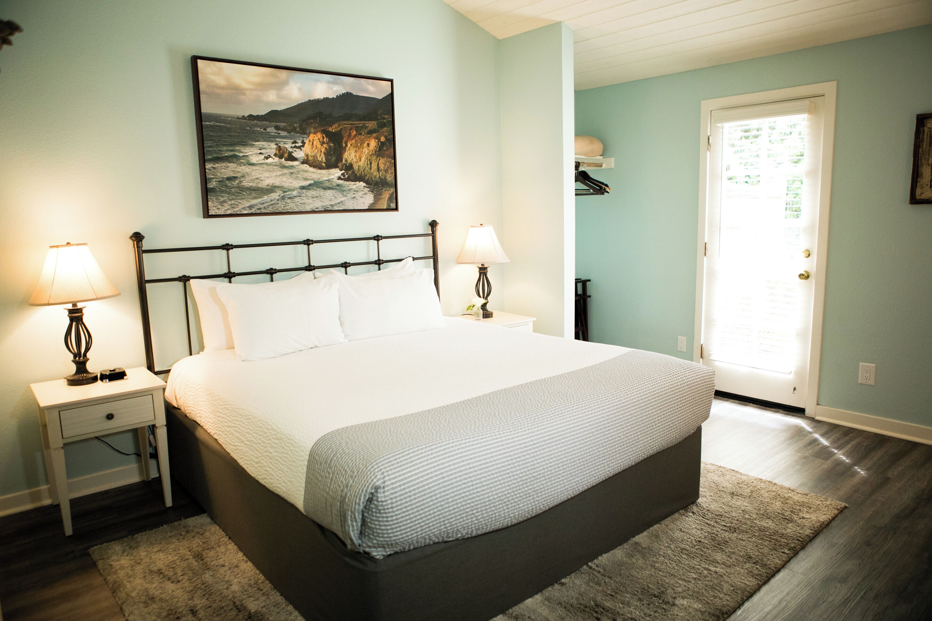 A bedroom with a bed, two bedside tables, a nightstand, a lamp, a painting, and a door.