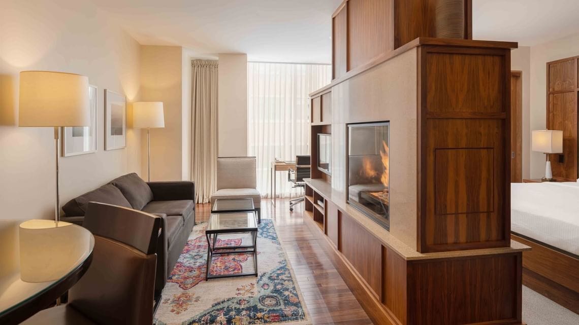 A hotel room with a sofa, armchair, TV, fireplace, and wooden cabinet.