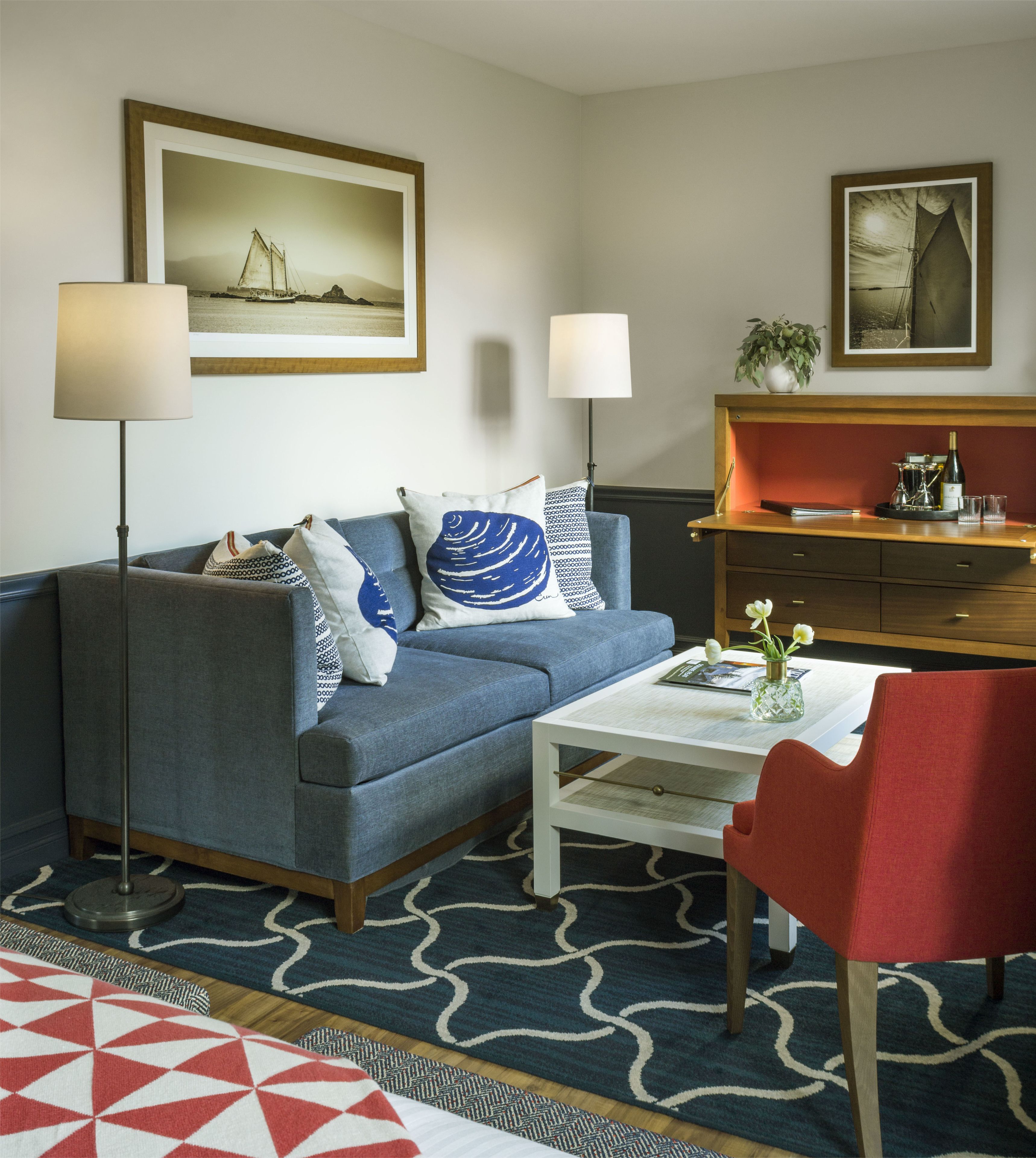 A living room with a blue sofa, a white coffee table, a red chair, and a wooden side table with a vase and glasses.