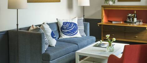 A living room with a blue sofa, a white coffee table, a red chair, and a wooden side table with a vase and glasses.