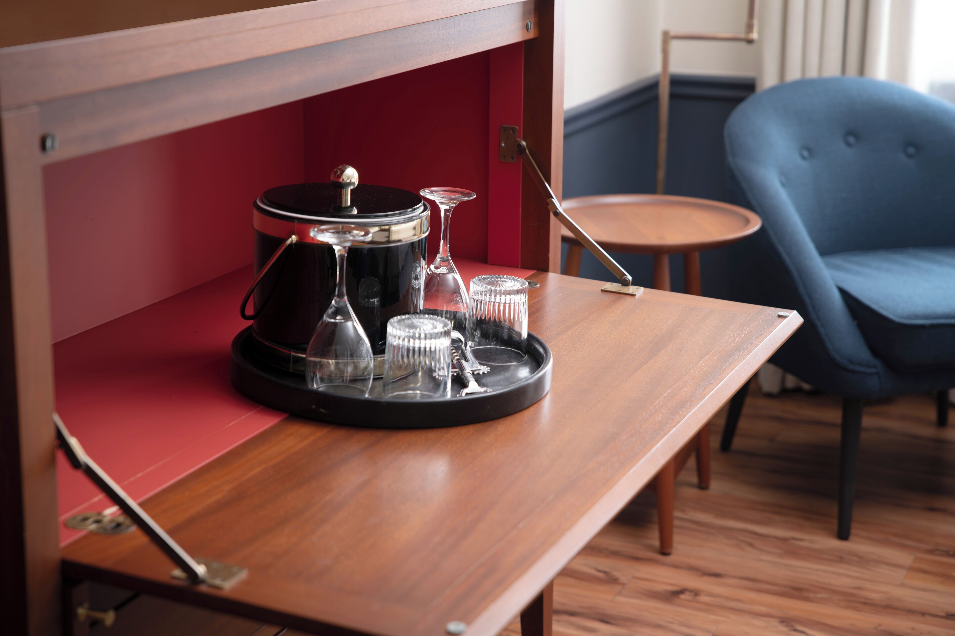 A wooden table with a coffee maker, glasses, and a tray in a room with a blue chair and a small round table.