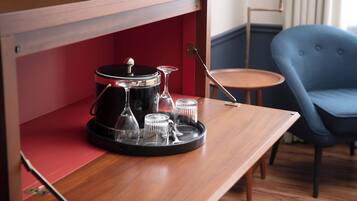 A wooden table with a coffee maker, glasses, and a tray in a room with a blue chair and a small round table.