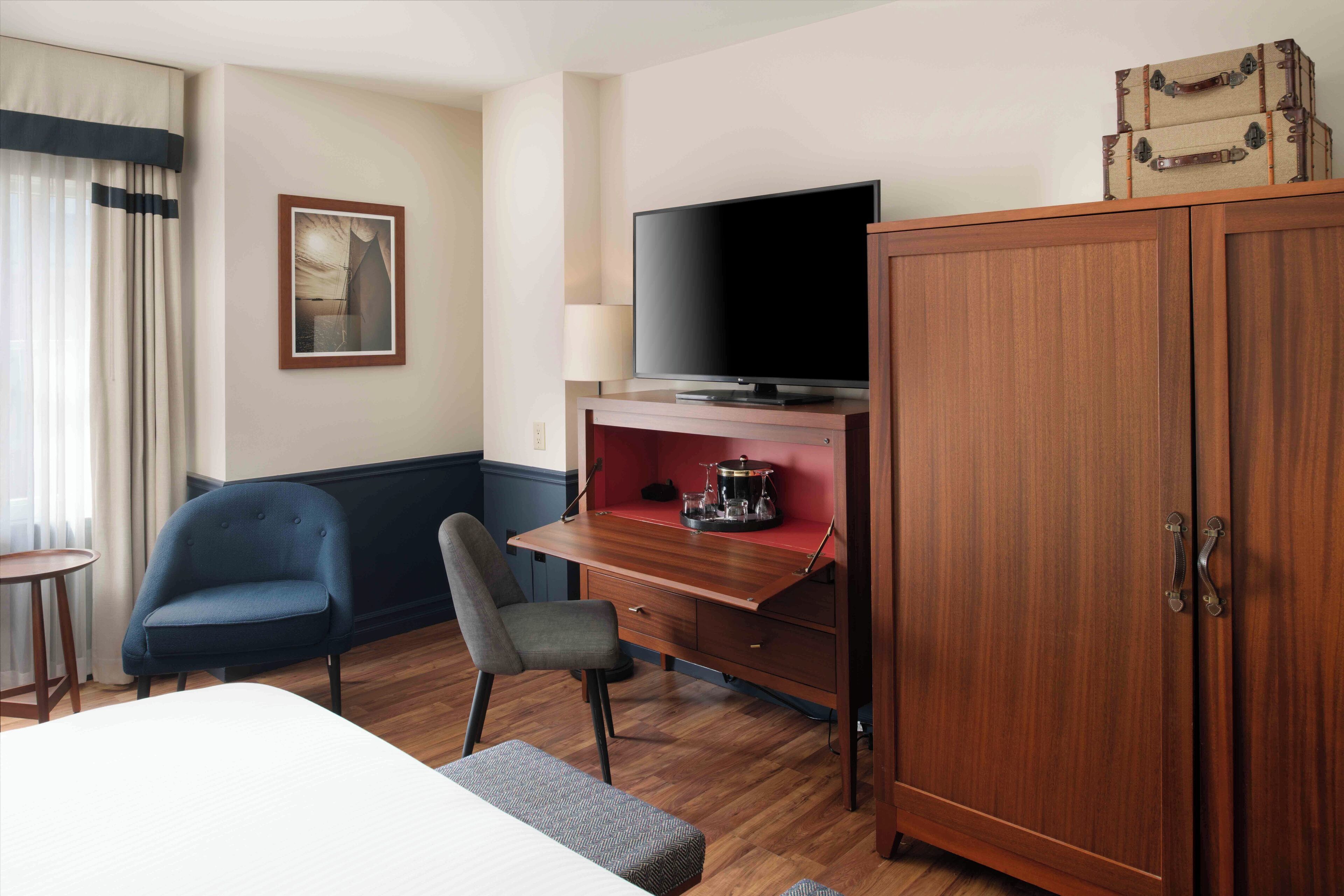 A hotel room with a bed, a desk with a television, a chair, a wardrobe, and a window with curtains.