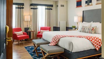 A hotel room with a large bed, two red armchairs, a small table with a lamp, and a view of the city through the window.
