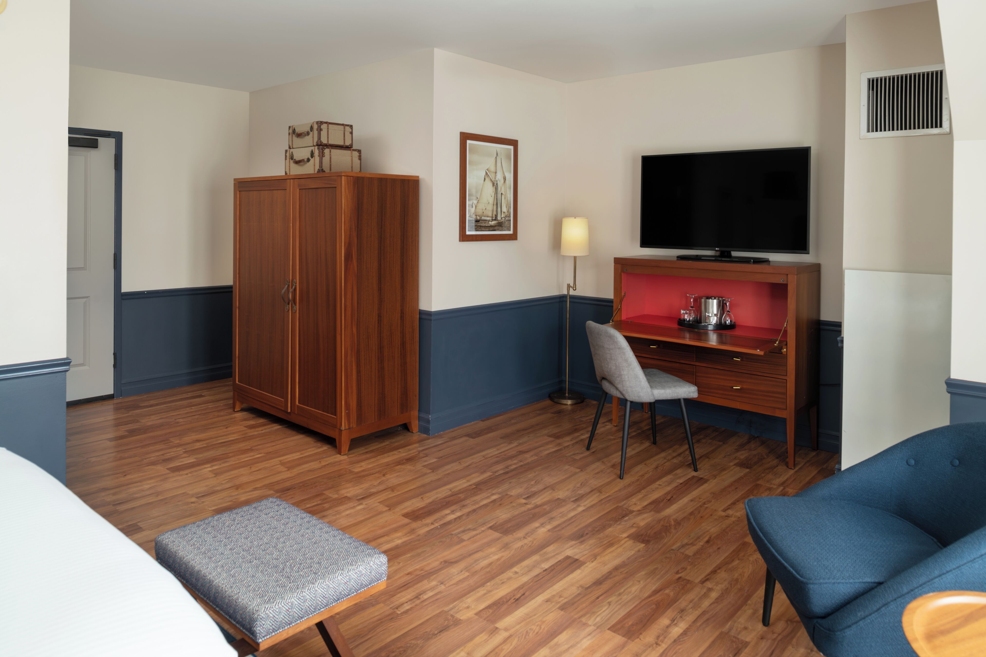 Ett hotellrum med trägolv, en säng, en blå fåtölj, ett skrivbord med en stol, en tv och en garderob.
