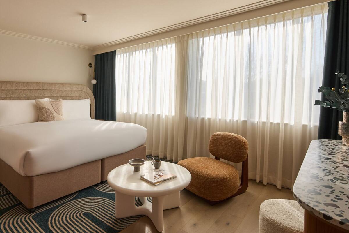 A hotel room with a bed, a chair, a small table, and a window with curtains.