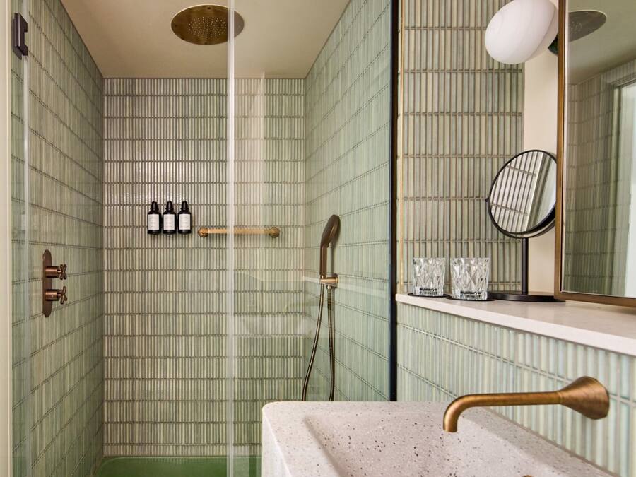 A bathroom with a shower, a sink with a brass faucet, a mirror, and a shelf with bottles and glasses.