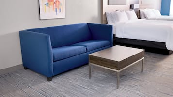A hotel room with a blue sofa, a wooden coffee table, a bed with white bedding, and a framed abstract artwork on the wall.