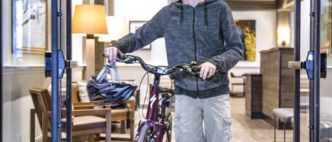 Una persona con bicicleta entrando en una habitación con piso de madera, una lámpara y cuadros en las paredes.