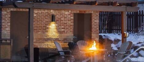 A heated outdoor pool area with lounge chairs and a fire feature, surrounded by a brick building and snow on the roof.