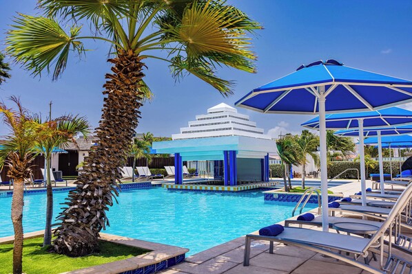 A pool area with lounge chairs and umbrellas, palm trees, and a building with a white facade.