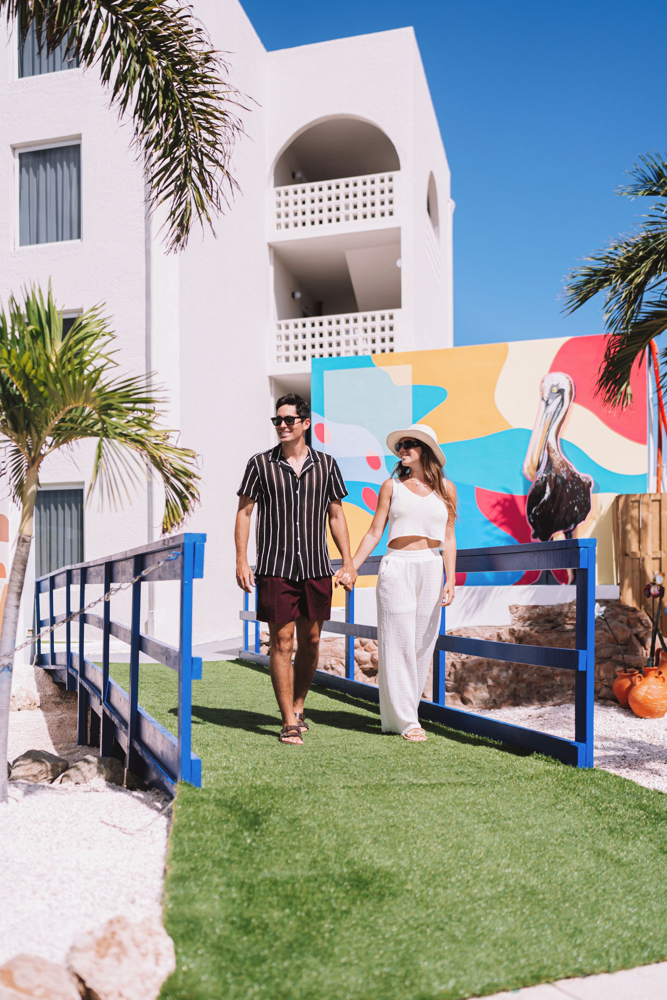 A couple walking on a pathway with a white building and a colorful mural in the background.