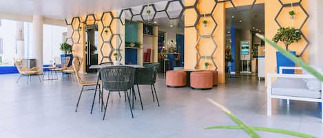 Un espacio interior moderno con paneles hexagonales en la pared, zonas de estar con mesas y sillas, y plantas en macetas.