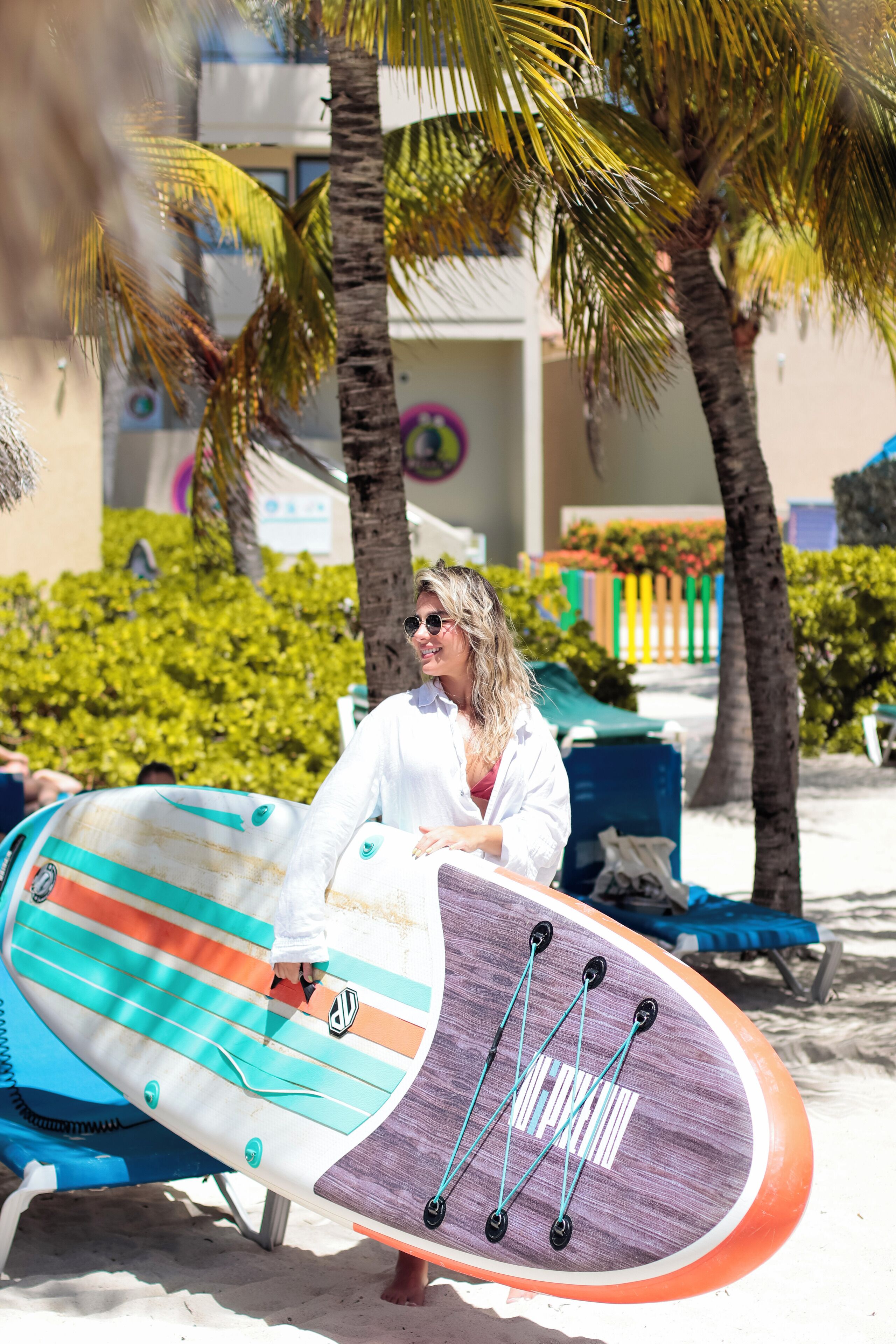 Une personne tenant une planche de surf sur une plage de sable, avec des palmiers et un bâtiment en arrière-plan.