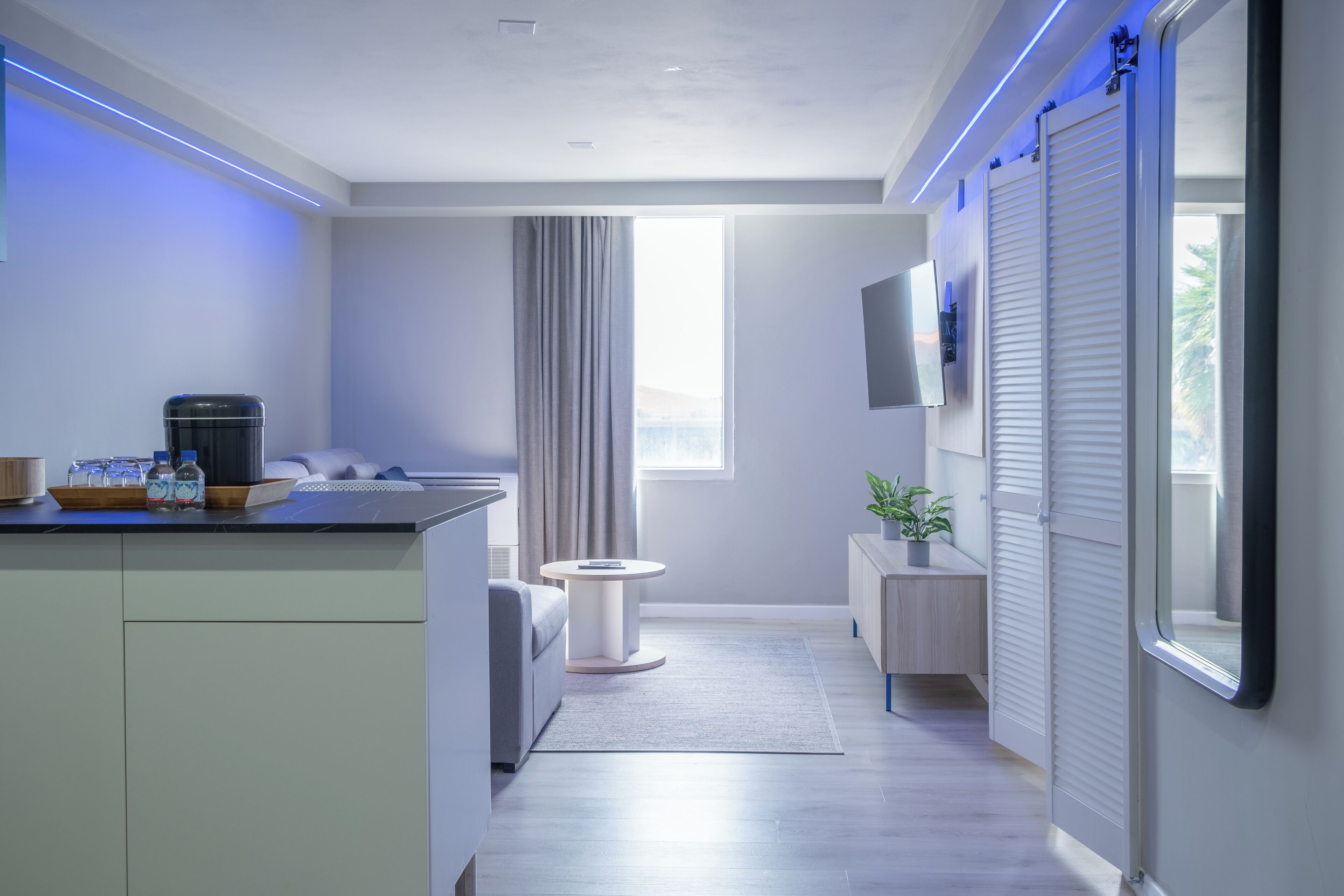 Une chambre d’hôtel moderne dotée d’une kitchenette, d’un téléviseur à écran plat fixé au mur, d’une grande fenêtre avec des rideaux et d’une porte coulissante.